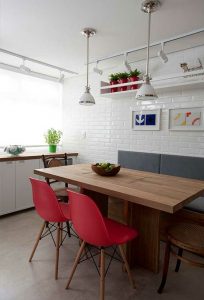 Sala de jantar com as paredes brancas, uma mesa de madeira no centro acompanhada de cadeiras vermelhas, luminárias brancas no teto e na prateleira vasos vermelhos. 