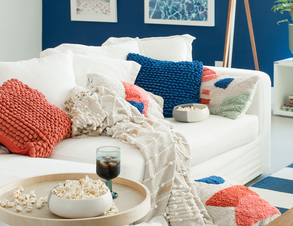 Sala de estar com uma parede azul e a outra branca, no centro um sofá branco acompanhado de uma manta na cor cru e diversas almofadas com textura, nas cores terracota e azul. Ao lado, uma mesa de apoio com um recipiente com pipoca e uma taça com bebida.