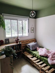 Quarto com uma pintura branca nas paredes e verde escura no telo. Uma cama com leçol verde e uma colcha rosa fica no canto direito do espaço e do outro lado, um móvel de madeira decorado com plantas e livros. 
