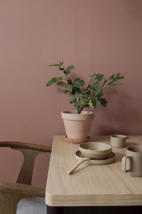Ambiente em tons rose e terrosos com uma mesa de madeira na direita da imagem, nela estão um vaso de cerâmica, pratos e canecas. 