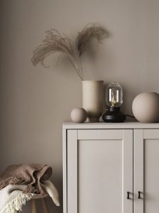 A foto mostra um armário na cor taupe, em sua parte superiro estão um vaso com plantas secas, uma luminária e dois vasos pequenos e redondos. Na lateral do armário há uma banco com duas mantas. 